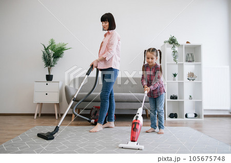 Young homemakers utilizing vacuum cleaners in living room 105675748
