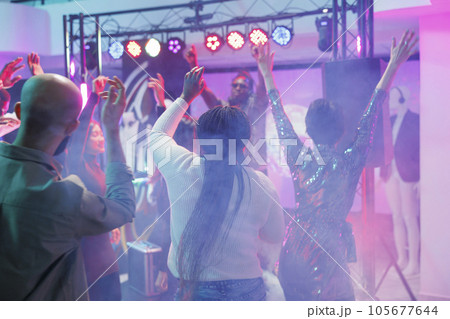 Energetic crowd dancing and enjoying discotheque on dancefloor with colorful spotlights. People jumping, having fun and moving to music beats with raised hands in nightclub 105677644