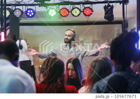 Man dj in headphones performing on nightclub stage at electronic music festival while people dancing. Young musician mixing tracks while clubbers crowd having fun on dancefloor 105677673