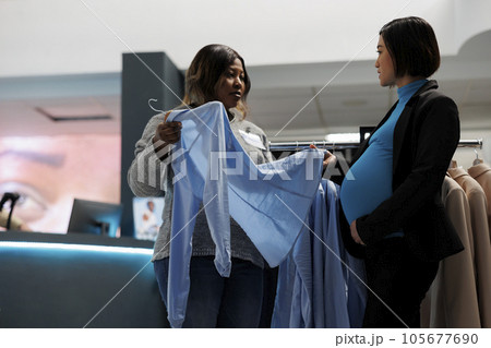 Shopping center assistant consulting pregnant woman in choosing maternity clothes. Boutique consultant showing client shirt on hanger, helping to select size and giving fashion advice Shopping center assistant consulting pregnant woman in choosing maternity clothes. Boutique consultant showing client shirt on hanger, helping to select size and giving fashion advice 105677690