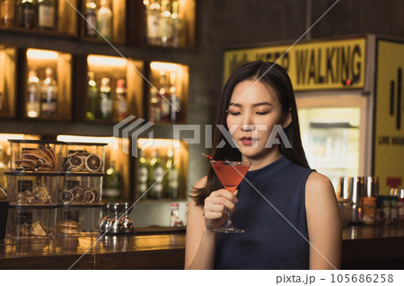 Asian woman drinking a cocktail at a bar at night. Asian woman drinking a cocktail at a bar at night. 105686258
