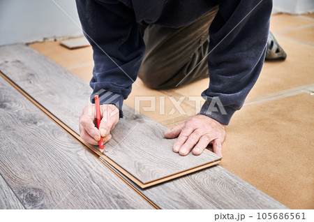 Close up of male construction worker with pencil in hand drawing line on laminate wooden panel. Man preparing laminate boards for floor installation in apartment under renovation. Close up of male construction worker with pencil in hand drawing line on laminate wooden panel. Man preparing laminate boards for floor installation in apartment under renovation. 105686561
