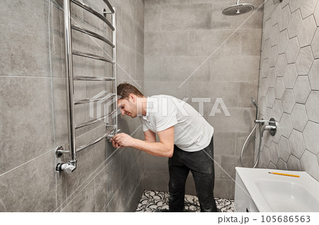 Man hanging electric towel rack dryer on the wall with ceramic tile in bathroom. Male worker installing bath towel heater in apartment under renovation. Concept of home renovation. Man hanging electric towel rack dryer on the wall with ceramic tile in bathroom. Male worker installing bath towel heater in apartment under renovation. Concept of home renovation. 105686563