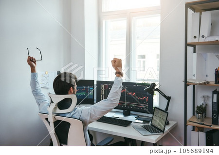 Winner Back view of stock trader with raised hands holding eyeglasses and feeling happy while looking at multiple computer screens with data and charts in his modern office. 105689194