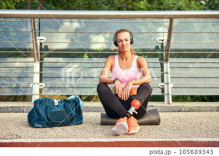 Confident in myself. Portrait of young woman in sports clothing and headphones with leg prosthesis sitting on the bridge and looking at camera Confident in myself. Portrait of young woman in sports clothing and headphones with leg prosthesis sitting on the bridge and looking at camera 105689343