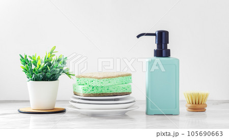 Cleaning sponge on plates with green dish soap bottle and washing brushes on white marble kitchen table 105690663