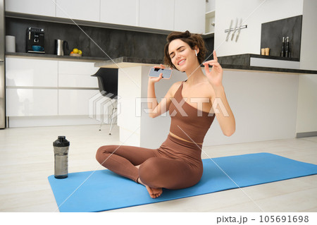 Active lifestyle and health. Happy young woman does workout, sits and dances with smartphone in hands, listens music in wireless earphones during fitness training session, yoga on floor mat 105691698