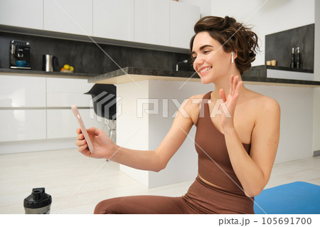 Young fitness girl at home, sport instructor in tracksuit waves hand at smartphone, sits on workout yoga mat, waves hand at mobile camera, says hi to online gym class, exercise remotely 105691700