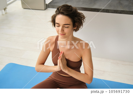 Young healthy beautiful woman in sports top and leggings practicing yoga at home, sitting in lotus position on yoga mat, meditating, smiling, relaxed, eyes closed, meditation concept 105691730