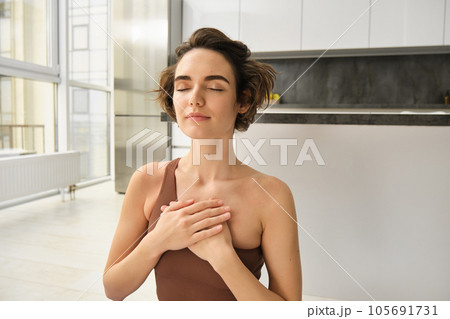 Portrait of young mindful woman, opens her chakra, doing yoga on rubber mat at home, closes eyes and smiles, feels peace inside her chest. 105691731