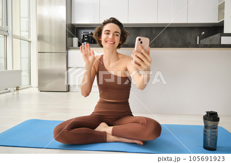 Smiling fitness girl, young athlete sits at home on yoga mat, waves hand at smartphone, joing online workout training session from home, says hello Smiling fitness girl, young athlete sits at home on yoga mat, waves hand at smartphone, joing online workout training session from home, says hello 105691923