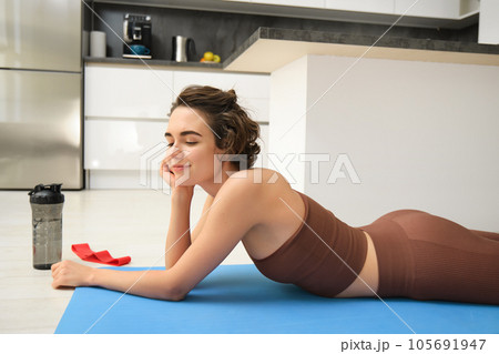 Sport and healthy lifestyle. Portrait of brunette sportswoman doing fitness in activewear, lying on rubber mat on floor, doing workout training in kitchen, drinking water from gym bottle Sport and healthy lifestyle. Portrait of brunette sportswoman doing fitness in activewear, lying on rubber mat on floor, doing workout training in kitchen, drinking water from gym bottle 105691947