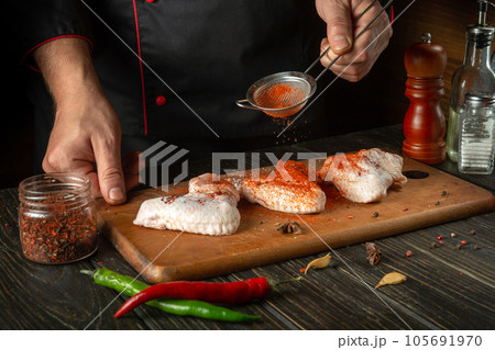 The chef or cook adds paprika to chicken wings. Grilled raw chicken wing in the kitchen of a restaurant or cafe on a kitchen table with vegetables and spices 105691970