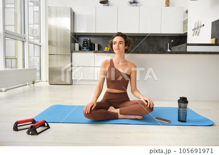 Portrait of young fitness woman, female athlete practice yoga from home, sitting in lotus pose with relaxed face, using rubber mat in kitchen for workout Portrait of young fitness woman, female athlete practice yoga from home, sitting in lotus pose with relaxed face, using rubber mat in kitchen for workout 105691971