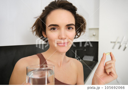 Portrait of sportswoman drinking water, taking vitamins, dietry supplements for healthy skin, having omega-3 pill, standing in her kitchen in workout clothing Portrait of sportswoman drinking water, taking vitamins, dietry supplements for healthy skin, having omega-3 pill, standing in her kitchen in workout clothing 105692066