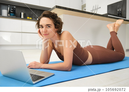 Portrait of beautiful sportswoman, fitness girl watching videos on laptop during workout, follow online pilates videos while exercise at home, wearing activewear in kitchen Portrait of beautiful sportswoman, fitness girl watching videos on laptop during workout, follow online pilates videos while exercise at home, wearing activewear in kitchen 105692072