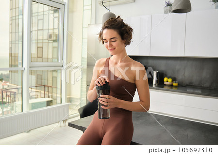 Sport and healthy lifestyle. Portrait of smiling fitness woman in activewear, standing near kitchen counter at home, drinking from water bottle, preparing for workout gym training Sport and healthy lifestyle. Portrait of smiling fitness woman in activewear, standing near kitchen counter at home, drinking from water bottle, preparing for workout gym training 105692310