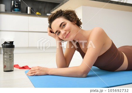 Sport and healthy lifestyle. Portrait of brunette sportswoman doing fitness in activewear, lying on rubber mat on floor, doing workout training in kitchen, drinking water from gym bottle Sport and healthy lifestyle. Portrait of brunette sportswoman doing fitness in activewear, lying on rubber mat on floor, doing workout training in kitchen, drinking water from gym bottle 105692732
