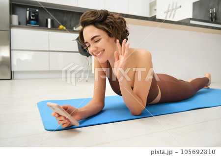 Portrait of happy fitness girl lying on rubber yoga mat, talking on mobile phone, saying hi at smartphone video chat, online training instructor connecting to gym session from home Portrait of happy fitness girl lying on rubber yoga mat, talking on mobile phone, saying hi at smartphone video chat, online training instructor connecting to gym session from home 105692810