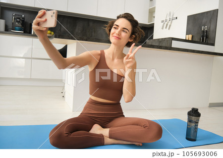 Portrait of young fitness woman, sitting at home on rubber mat, records video on smartphone, doing workout vlog, making live online training session from home Portrait of young fitness woman, sitting at home on rubber mat, records video on smartphone, doing workout vlog, making live online training session from home 105693006