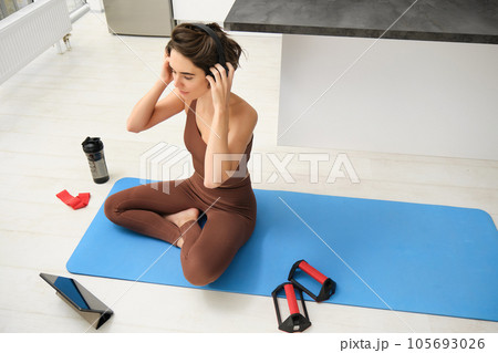 Smiling fitness girl in headphones, watches pilates, gym instructor online video, workout at home on yoga rubber mat, uses plank equipment and resistance band Smiling fitness girl in headphones, watches pilates, gym instructor online video, workout at home on yoga rubber mat, uses plank equipment and resistance band 105693026