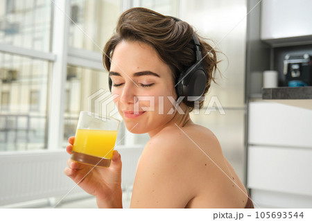 Sport and healthy lifestyle. Portrait of happy sporty girl, fitness woman in headphones, rewinds at home, drinks orange juice with happy smile after workout 105693544