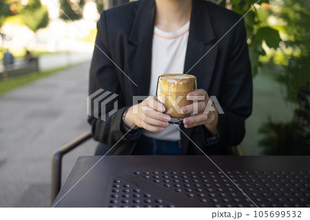 Close up of young female in business casual wear drinking cup of coffee while sitting at small table and enjoying time in street cafe. Close up of young female in business casual wear drinking cup of coffee while sitting at small table and enjoying time in street cafe. 105699532