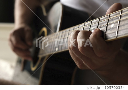 Playing the guitar. Strumming acoustic guitar. Musician plays music. Man fingers holding mediator. Man hand playing guitar neck in dark room. Unrecognizable person rehearsing, fretboard close-up. 105700324
