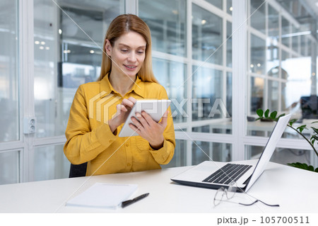 Mature woman boss inside the office at the desk uses a laptop and a tablet computer, a businesswoman is satisfied with the financial results and browses online pages on the Internet. Mature woman boss inside the office at the desk uses a laptop and a tablet computer, a businesswoman is satisfied with the financial results and browses online pages on the Internet. 105700511