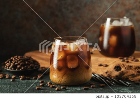 Ice coffee in a tall glass with cream poured over, ice cubes and beans on a dark concrete table. Ice coffee in a tall glass with cream poured over, ice cubes and beans on a dark concrete table. 105704359