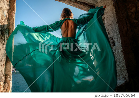 Rear view of a happy blonde woman in a long mint dress posing against the backdrop of the sea in an old building with columns. Girl in nature against the blue sky. Rear view of a happy blonde woman in a long mint dress posing against the backdrop of the sea in an old building with columns. Girl in nature against the blue sky. 105705664