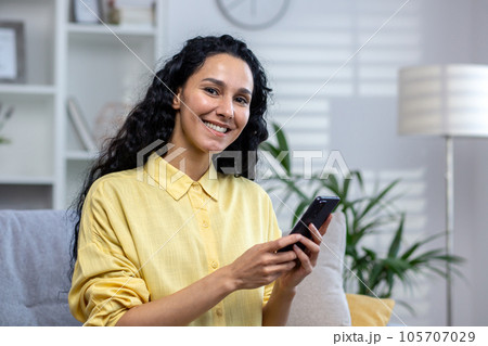 Portrait of a young beautiful hispanic woman at home, smiling and looking at the camera, holding a phone in her hands, using an application on a smartphone, browsing the Internet, typing a message. 105707029
