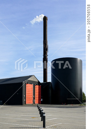 Modern factory with a tall rusty chimney from which comes out white steam on blue sky background Modern factory with a tall rusty chimney from which comes out white steam on blue sky background 105708558