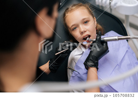High-angle view portrait of cute little child girl sitting in stomatology seat with open mouth while unrecognizable female orthodontist examining teeth, with dental tool in dentistry clinic. High-angle view portrait of cute little child girl sitting in stomatology seat with open mouth while unrecognizable female orthodontist examining teeth, with dental tool in dentistry clinic. 105708832