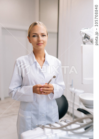Vertical portrait of female dental doctor holding working equipment standing by chair and looking at camera in dentistry clinic with modern white interior. Concept of stomatology, dentistry. Vertical portrait of female dental doctor holding working equipment standing by chair and looking at camera in dentistry clinic with modern white interior. Concept of stomatology, dentistry. 105708840