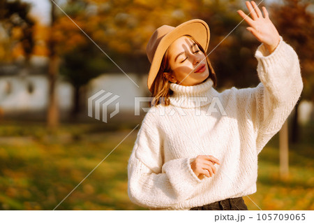 Autumn woman. Young cheerful woman in white sweater and hat walks in the nature park on a sunset. 105709065