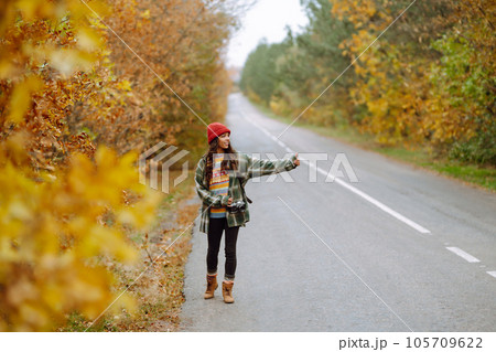 Young female hitchhiker by roadside among autumn forest during fall season. Travel woman hitchhiking 105709622