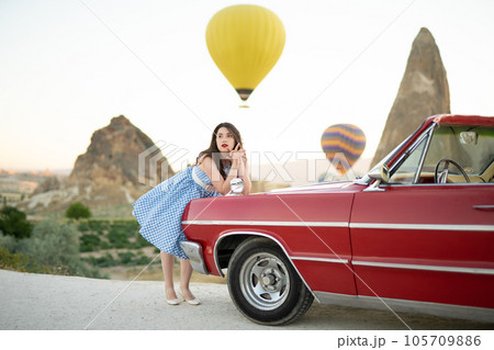 beautiful girl in retro style posing near a vintage red cabriolet car on background of balloons in Cappadocia. beautiful girl in retro style posing near a vintage red cabriolet car on background of balloons in Cappadocia. 105709886