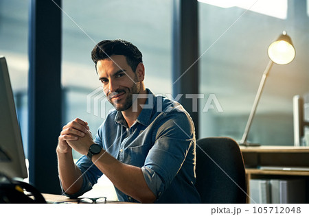Portrait of a young businessman on a computer during a late night at work. Confident hardworking man at a corporate office desk working overtime, dedicated and committed alone at the workplace. Portrait of a young businessman on a computer during a late night at work. Confident hardworking man at a corporate office desk working overtime, dedicated and committed alone at the workplace. 105712048