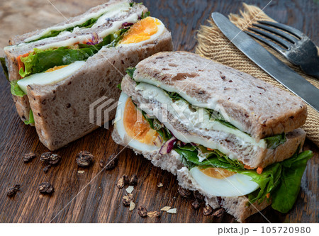 Sandwich stuffed with chicken breast, cheese, boiled eggs and fresh vegetables serve with fork and knife on wooden plate. 105720980
