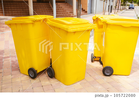 Four yellow plastic trash cans stand on a city street near apartment buildings in a residential neighborhood. Urban Scene. Ecology, cleanliness concept. Gathering rubbish. Wheelie trash bins outdoors. 105722080