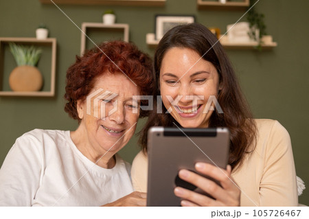 Portrait of a modern happy grandmother and granddaughter sitting in the living room during the day and looking cheerfully at the tablet Portrait of a modern happy grandmother and granddaughter sitting in the living room during the day and looking cheerfully at the tablet 105726467
