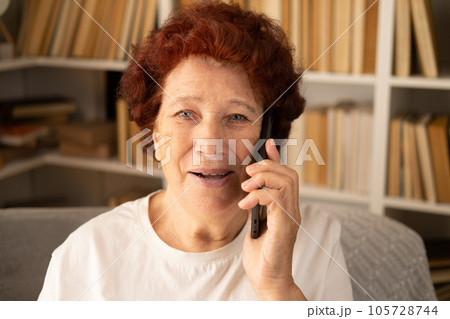 Close-up portrait of a modern elderly woman who sits in a chair against the background of a bookcase, talking on the phone and looking at the camera. Close-up portrait of a modern elderly woman who sits in a chair against the background of a bookcase, talking on the phone and looking at the camera. 105728744