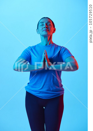 Beautiful mature woman standing in yoga pose, relaxing body and mind against blue studio background in neon light Beautiful mature woman standing in yoga pose, relaxing body and mind against blue studio background in neon light 105729510