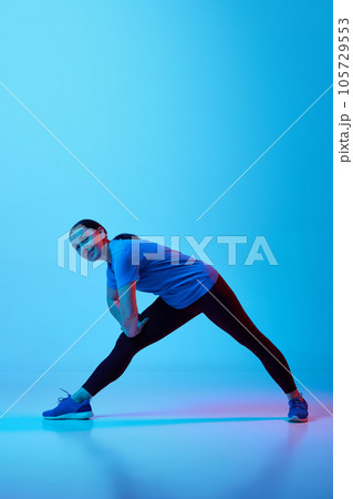 Reduction of knee pain. Mature slim woman in black sportswear stretching legs, training against blue studio background in neon light Reduction of knee pain. Mature slim woman in black sportswear stretching legs, training against blue studio background in neon light 105729553