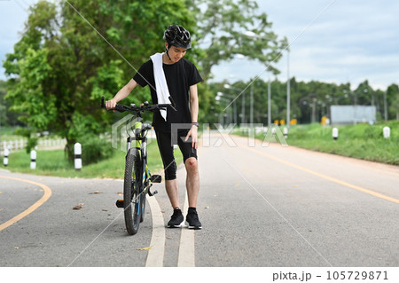 Shot of male cyclist with injured knee carry bicycle walking in park after an accident Shot of male cyclist with injured knee carry bicycle walking in park after an accident 105729871