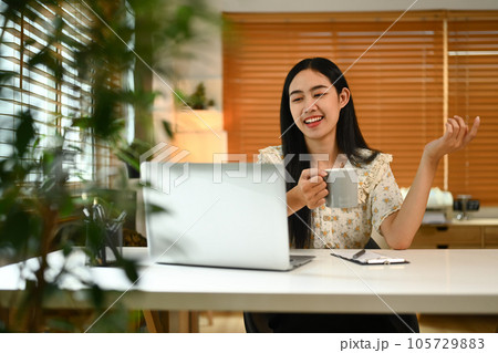 Nice, friendly young woman drinking coffee and talking on video call with friends via laptop at home Nice, friendly young woman drinking coffee and talking on video call with friends via laptop at home 105729883