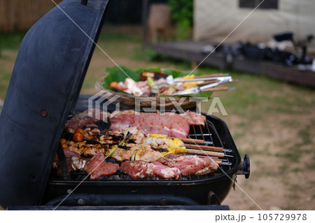 Meat skewers and vegetables on a BBQ grill near camping tent outdoors in evening. 105729978