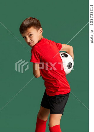 Future football player. Little boy, child in sportswear posing, playing with ball against green studio background 105735811