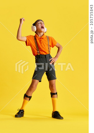 Full-length of little boy, child in stylish clothes listening to music in headphones and singing against yellow studio background 105735843
